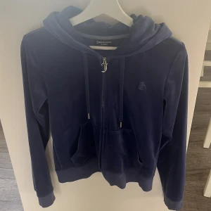 Blå velour hoodie från Juicy Couture - Mörkblå hoodie från Juicy Couture i mjuk velour med dragkedja och huva. Klassisk JC-logga på bröstet och snygg J-detalj på dragkedjan. Perfekt för en chill och trendig look. Fickor framtill och långa ärmar. 