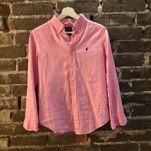 Rosa skjorta från Ralph Lauren - Klassisk rosa skjorta från Ralph Lauren med button-down krage och broderad marinblå logga på bröstet. Skjortan har långa ärmar, knappar framtill och är tillverkad i mjuk bomull och linne. Perfekt för en stilren och avslappnad look.