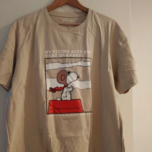 Beige Peanuts Snoopy t-shirt Uniqlo - Beige t-shirt från Uniqlo UT med Peanuts-tryck på framsidan där Snoopy är utklädd till flygare. Skön bomullskvalitet och klassisk passform med rund hals och korta ärmar. Perfekt för dig som gillar coola och lekfulla prints.