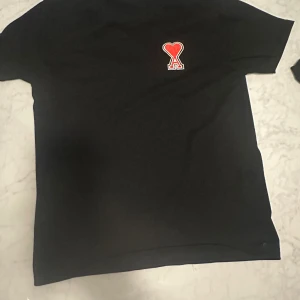 Svart t-shirt från Ami Paris med hjärtdetalj - Svart t-shirt från Ami Paris med klassisk passform och korta ärmar. På bröstet sitter det ikoniska röda Ami de Coeur-hjärtat broderat. T-shirten är gjord i mjuk bomull och har en stilren look som passar till allt.