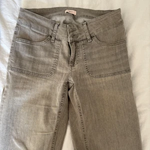 Grå bootcut jeans från Nelly - Snygga grå jeans från Nelly med bootcut passform och låg midja. Jeansen har dubbla knappar i midjan, stora framfickor och bakfickor med lock och knapp. Materialet är klassisk denim och färgen är en cool, ljusgrå ton.