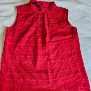 Röd ärmlös blus från NSF - Säljer en snygg röd blus från NSF i storlek L. Blusen är ärmlös med hög krage och har ett diskret randigt mönster i tyget. Materialet känns lätt och luftigt, perfekt för varmare dagar. Passformen är normal och toppen har en stilren look.