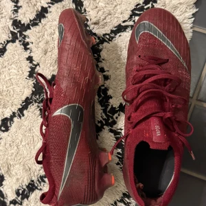 Röda Nike Vapor 360 fotbollsskor - Säljer ett par röda Nike Vapor 360 fotbollsskor med silverfärgade swoosh-detaljer. Skorna har snörning och är tillverkade i syntetmaterial med mönstrad ovandel och dobbar för gräsplan. Perfekta för dig som vill sticka ut på planen.