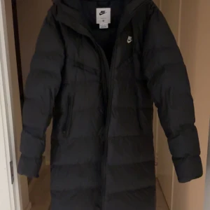 Svart lång dunjacka från Nike XS - Nike dunjacka i svart med huva och Storm-FIT-teknologi. Lång modell med quiltad design, dragkedja framtill och sidofickor med dragkedja. Perfekt för kalla dagar, stilren och sportig look med Nike-logga på bröstet.