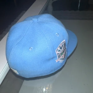 Blå LA Dodgers keps med brodyr - Säljer en blå keps från New Era med LA Dodgers-logga i ljusrosa framtill och broderade palmer i grönt. Kepsen har platt skärm och broderad detalj på sidan. Perfekt för dig som vill ha något unikt 