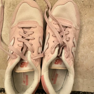 Ljusrosa New Balance sneakers - Säljer ett par snygga ljusrosa New Balance sneakers med vit N-logga på sidan. Skorna har snörning, platt sula och rund tå. Ovandelen är i mesh och mocka, med detaljer i syntet. Perfekta för dig som gillar sportig stil och pastellfärger.