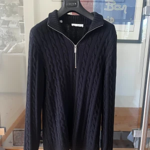 Svart kabelstickad tröja Selected Homme - Svart kabelstickad tröja från Selected Homme med halv dragkedja och hög krage. Tröjan har lång ärm och ribbade muddar, perfekt för lager-på-lager under kyliga dagar. Klassisk och stilren look som funkar till det mesta.