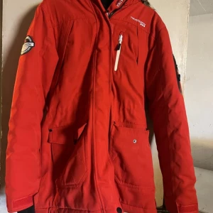 Röd parkajacka från True North - Säljer en röd parkajacka från True North med flera fickor, dragkedja och snygga detaljer. Jackan har lång ärm, hög krage och är perfekt för kalla dagar. Materialet är slitstark polyester och designen är clean med logga på bröstet och ärmen.
