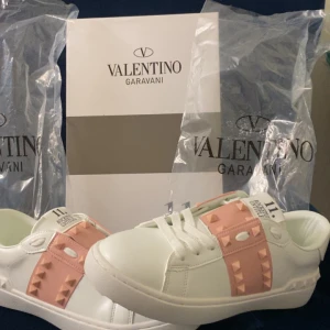 Valentino Rockstud sneakers vit/rosa - Snygga Valentino Garavani Rockstud sneakers i vitt skinn med ljusrosa detaljer och ikoniska nitar. Skorna har rund tå, platt sula och snörning framtill. Perfekta för dig som vill ha en lyxig och trendig look med en touch av rosa.