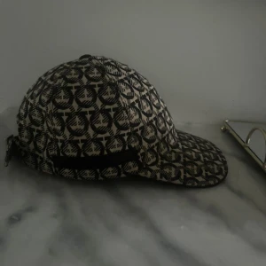Ferregamo keps - Säljer en keps i beige och svart med ett grafiskt, återkommande mönster över hela kepsen. Justerbar rem baktill med metallspänne. Kepsen har böjd skärm och klassisk sexpanelsdesign. Perfekt accessoar för en trendig look.