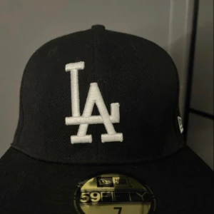 Svart LA Dodgers keps New Era 59FIFTY - Svart keps från New Era 59FIFTY med LA Dodgers broderad logga i vitt framtill och MLB-logga bak. Klassisk rak skärm och strukturerad passform. Tillverkad i polyester och ullblandning. Perfekt för dig som gillar sportig streetstyle.