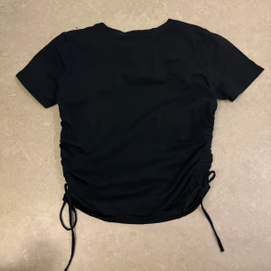 Svart topp med snörning från Zara - Svart kortärmad topp från Zara med snörning i sidorna som ger en snygg rynkad effekt. T-shirten har en klassisk rund halsringning och är tillverkad i mjukt material som känns skönt mot huden. Perfekt för att skapa en trendig look.