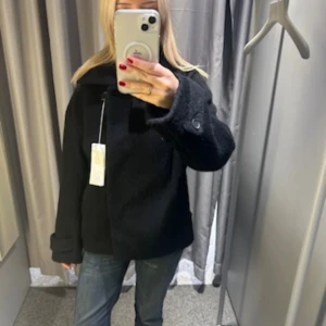 Svart skepparkavaj med stora knappar - Säljer en svart kappa från arket med oversized passform och stora knappar vid ärmslut och hals. Jackan har hög krage som kan knäppas upp och är tillverkad i ett ulligt material. Perfekt för dig som vill ha en stilren och mysig jacka till höst och vinter. Saknas en knapp i mitten på insidan men inget som känns eller syns.