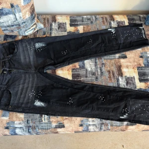 Svarta jeans med vita detaljer - Säljer ett par svarta jeans med coola vita stänk och prickdetaljer. Byxorna har raka ben och klassisk femficksdesign, med extra fickor baktill. Materialet är jeans och färgen är mörk, nästan tvättad svart. Perfekta för dig som vill sticka ut lite extra.