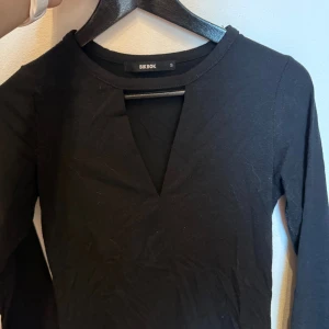 Svart långärmad topp från BikBok - Svart långärmad topp från BikBok med snygg v-ringning framtill. Enkel och stilren design i mjukt bomullsmaterial, perfekt att matcha med jeans eller kjol. Toppen har en figurnära passform och är superenkel att styla.