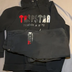 Svart Trapstar hoodie med byxor - Svart hoodie från Trapstar med färgstarkt tryck i rött, grått och vitt på bröstet. Klassisk känguruficka framtill, snörning i huvan och mjukt material som känns skönt mot huden. Perfekt för dig som gillar streetwear och vill sticka ut.