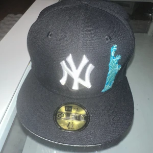 New Era Yankees svart keps  - Svart New Era 59FIFTY keps med New York Yankees-logga framtill i vitt, Frihetsgudinnan broderad i blått på sidan och ett rött äpple broderat baktill. Klassisk flat peak och broderade ventilationshål. Perfekt för dig som gillar streetwear och sportig stil.