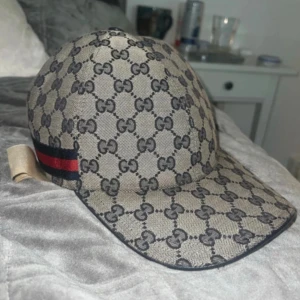 Gucci keps med GG-mönster och röd-blå detalj - Snygg beige keps från Gucci med klassiskt GG-monogram över hela ytan. Kepsen har en röd och blå rand på sidan och justerbar rem baktill. Materialet är bomull och kepsen har en rundad form med böjd skärm. Perfekt för dig som vill ha en lyxig streetstyle-look.