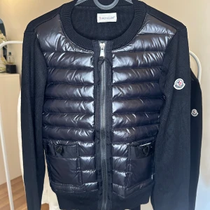 Moncler Cardigan  - Svart cardigan jacka från Moncler med quiltad, glansig front och stickade ärmar. Jackan har dragkedja framtill, två fickor med knappdetaljer och Moncler-logga på ärmen. Perfekt för dig som gillar en sportig och stilren look.