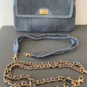 Blå axelväska i äkta skinn - Snygg blå axelväska i äkta skinn med både mocka och släta detaljer. Väskan har två utbytbara axelband, ett flätat i skinn och ett med guldfärgad kedja. Stängs med vridlås i guld och har flera fack inuti samt dragkedja på baksidan. Mått Bredd: ca 31 cm Höjd: ca 20 cm  Kommer från ett djur och rökfritt hem. 