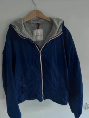 Blå vindjacka med huva från Moncler - Snygg blå vindjacka från Moncler med grå huva och dragkedja framtill. Jackan har Moncler-logga på ärmen och vita, röda detaljer längs dragkedjan. Perfekt för vår och höst, tillverkad i lätt polyester och har långa ärmar.