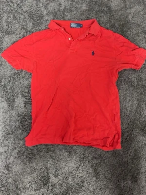 Ralph Lauren piké  - Klassisk röd pikétröja från Polo Ralph Lauren med korta ärmar och krage. Tröjan har två knappar vid halsen och den ikoniska blå loggan broderad på bröstet. Perfekt för en stilren och avslappnad look.