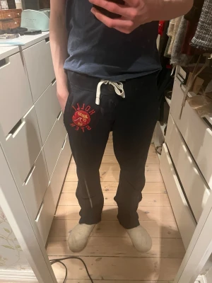 Mörkblåa mjukisbyxor från Hollister - Mörkblåa mjukisbyxor från Hollister med vit snörning i midjan och röd logga på vänster lår. Sköna och avslappnade med normal passform, perfekta för chill dagar. Tillverkade i mjukt bomullsmaterial.