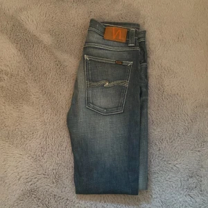 Blå jeans från Nudie Jeans, 34/34 - Snygga blå jeans från Nudie Jeans med klassisk femficksdesign och coola kontrastsömmar på bakfickorna. Modellen har en rak passform och subtila slitningar framtill och baktill för en avslappnad vibe. 