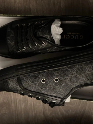 Svarta Gucci sneakers med monogram - Snygga svarta sneakers från Gucci med diskret monogrammönster över hela skon. Skorna har snörning, svarta detaljer i skinn och en platt sula. Perfekta för dig som vill ha en exklusiv och stilren look.