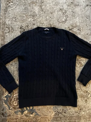 Mörkblå kabelstickad tröja GANT - Klassisk mörkblå kabelstickad tröja från GANT med rund halsringning och diskret logga broderad på bröstet. Tröjan har ribbade muddar vid ärmslut och nederkant, perfekt för lager på lager och en stilren look.