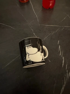 Svart Muminmugg  Arabia Finland - Cool svart mugg med Muminpappa-motiv från Arabia Finland. Muggen är i keramik och har ett vitt tryck av Muminpappan med hatt och käpp. Perfekt för te eller kaffe och en riktig samlarpryl för Muminfans.