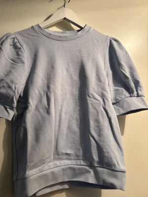 Ljusblå puffärmad topp - Säljer en ljusblå topp med puffärmar och rund hals. Toppen har korta ärmar med breda muddar och är gjord i mjuk bomull. Enkel och stilren design som passar perfekt till jeans eller kjol.