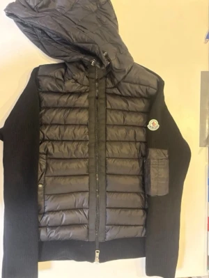Svart cardigan jacka från Moncler - Svart cardigan jacka från Moncler med quiltad framsida och huva. Jackan har stickade ärmar och sidor, dragkedja framtill och en ficka på ärmen. Ikonisk Moncler-logga på vänster ärm. Perfekt för lager-på-lager och snygg streetstil.
