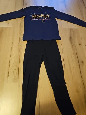 Harry Potter pyjamas set blå/svart - Mysig pyjamas med långärmad blå tröja och svarta byxor. Tröjan har Harry Potter-tryck med text och motiv av Nimbus 2000. Byxorna är enkla och svarta med resår i midjan. Perfekt för alla Potterheads som vill chilla hemma. Tröjan stl 146-152 ,byxorna är matchande men är inte med i det orginella stället och är stl 134-140.