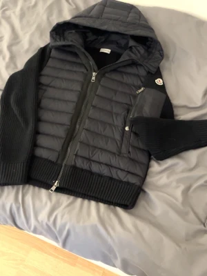 Svart teddyjacka från Moncler - Snygg svart teddyjacka från Moncler med quiltad front, stickade ärmar och huva. Jackan har dragkedja framtill och en ficka med Moncler-logga på ärmen. Perfekt för dig som vill ha en stilren och bekväm jacka.