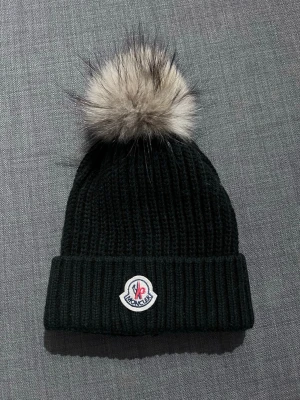 Svart Moncler mössa med pälsboll - Snygg svart ribbstickad mössa från Moncler med stor beige pälsboll på toppen och vikt kant. Klassisk Moncler-logga framtill. Perfekt accessoar för kalla dagar och ger en trendig look till din outfit.