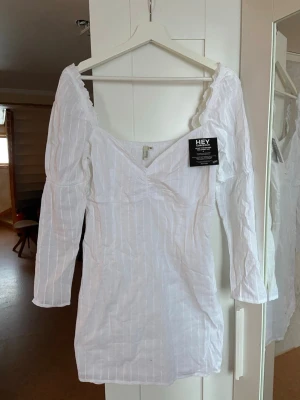 Vit kort klänning från Nelly - Säljer en vit kort omlottklänning från Nelly med lång ärm och v-ringad offshoulder-modell. Klänningen har diskreta ränder och är tillverkad i ett lätt bomullstyg, perfekt för varmare dagar. Säljer den då den är för stor för mig, aldrig använd och lappen kvar. 🩵