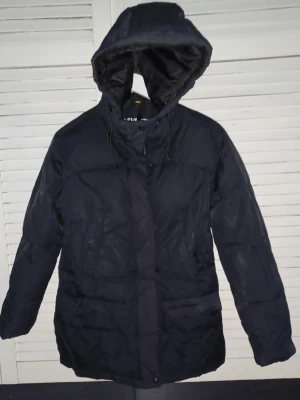 Svart dunjacka med huva - Mörk, lång svart dunjacka med huva och dragsko. Jackan har en quiltad design och stängs med dragkedja och knappar framtill. Perfekt för kalla vinterdagar och har en klassisk, enkel look som passar till det mesta.