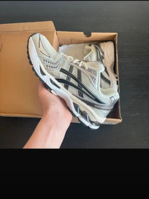 Asics Gel-Kayano 14 beige/silver - Säljer ett par Asics Gel-Kayano 14 sneakers i beige och silver med svarta detaljer. Skorna har en sportig design med mesh och syntetmaterial, samt en chunky sula för extra komfort. Perfekta för dig som gillar retrovibbar och streetstyle.