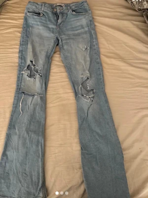 Ljusblå bootcut jeans från Zara - Säljer ett par ljusblå bootcut jeans från Zara med slitna detaljer och stora hål på båda benen. Jeansen har klassisk femficksdesign och är tillverkade i bomull. Perfekta för en avslappnad och trendig look.