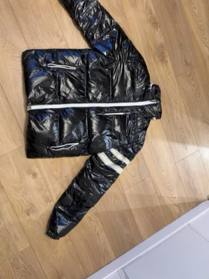 Svart Jacka Moncler; Pris kan diskuteras - Snygg svart dunjacka från Moncler med glansig finish och vita detaljer. Jackan har vita dragkedjor, en vit rand på ena ärmen och quiltad design. Insidan är vit och jackan har flera fickor med dragkedja. Perfekt för kalla dagar.