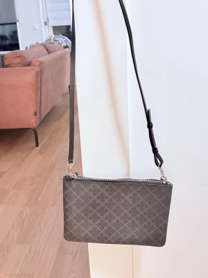 Malene Birger Kuvertväska - Snygg grå axelväska med rutigt mönster och justerbar axelrem. Väskan har en stilren rektangulär form och stängs med dragkedja upptill. Perfekt för att bära det viktigaste och passar till många olika outfits.