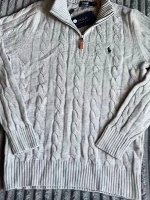 Grå stickad tröja Polo Ralph Lauren - Klassisk grå stickad tröja från Polo Ralph Lauren med kabelstickat mönster och blå broderad logga på bröstet. Rund halsringning och långa ärmar. Perfekt till jeans  för en clean och tidlös look.När köparen beställer skickar vi varan på samma dag eller dagen där på, Varan är aldrig använd. 
