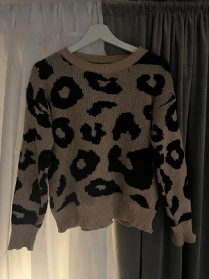 Beige leopardmönstrad stickad tröja - Säljer en ljusbrun/beige stickad tröja med svart leopardmönster. Tröjan har rund halsringning, ribbade muddar och lång ärm. Perfekt för dig som vill ha en trendig och mysig look med lite edge.