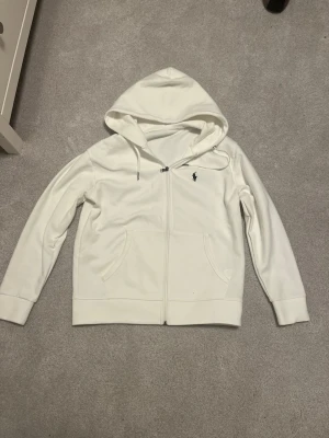Vit hoodie från Polo Ralph Lauren - Säljer en stilren vit hoodie från Polo Ralph Lauren med dragkedja framtill och två fickor. Klassisk logga broderad på bröstet och justerbar huva med snören. Tillverkad i mjukt bomullsmaterial som känns skönt mot huden.