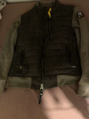 Mörkgrön dunjacka från Parajumpers - Snygg mörkgrön dunjacka från Parajumpers med quiltad front och ärmar i matchande färg. Jackan har dragkedja framtill, två fickor med dragkedja och en gul detalj vid halsen. Perfekt för kyliga dagar.