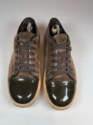 Bruna Lanvin skor med lackad tå - Snygga bruna sneakers i mocka med mörkbruna snören och en glansig, lackad tå. Skorna har beige sula och klassisk rund tå. Perfekta för dig som gillar en stilren men unik look.