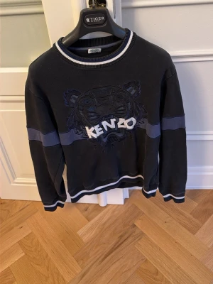 Svart Kenzo sweatshirt med tigertryck - Cool svart sweatshirt från Kenzo med det ikoniska tigerbroderiet och loggan framtill. Tröjan har vita och blå detaljer vid krage, ärmslut och nederkant samt en blå rand över bröstet och armarna. Perfekt för en trendig streetwear-look.