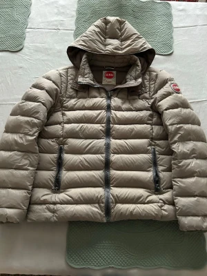 Beige dunjacka med huva från Colmar - Snygg beige dunjacka från Colmar med huva och två dragkedjeförsedda fickor framtill. Jackan har quiltad design och Colmar-logga på ärmen. Perfekt för kyliga dagar och har en modern, puffig look. Storlek 50/M