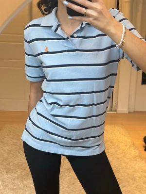 Blå- och vitrandig piké från Ralph Lauren - Snygg pikétröja från Ralph Lauren i ljusblått med vita och mörkblå horisontella ränder. Klassisk krage och korta ärmar, samt den ikoniska orangea logotypen broderad på bröstet. Perfekt till jeans eller shorts för en avslappnad look.